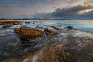 Seascape sunrise long exposure with flowing water around large rocks and light pink and blue sky