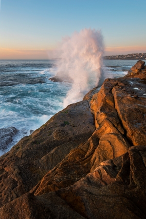 Sunrise seascape with unrest sea and blue water and with sky lit orange rocks and big crashing wave