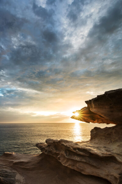 The sun rising over the rugged cliffs at Cape Solander, casting a warm golden light.