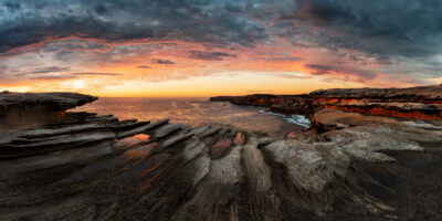 Panoramic view of Cape Solander at sunrise with fiery sky and textured rocks.