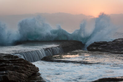A powerful wave crashes over rocks at Clovelly Beach during sunrise, creating a dramatic display of nature's force.