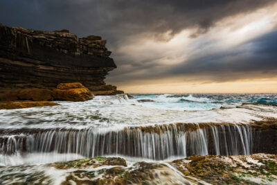 Stormy sunrise at Clovelly Beach showing water flowing over rocky ledges into the abyss below, under a tumultuous sky.