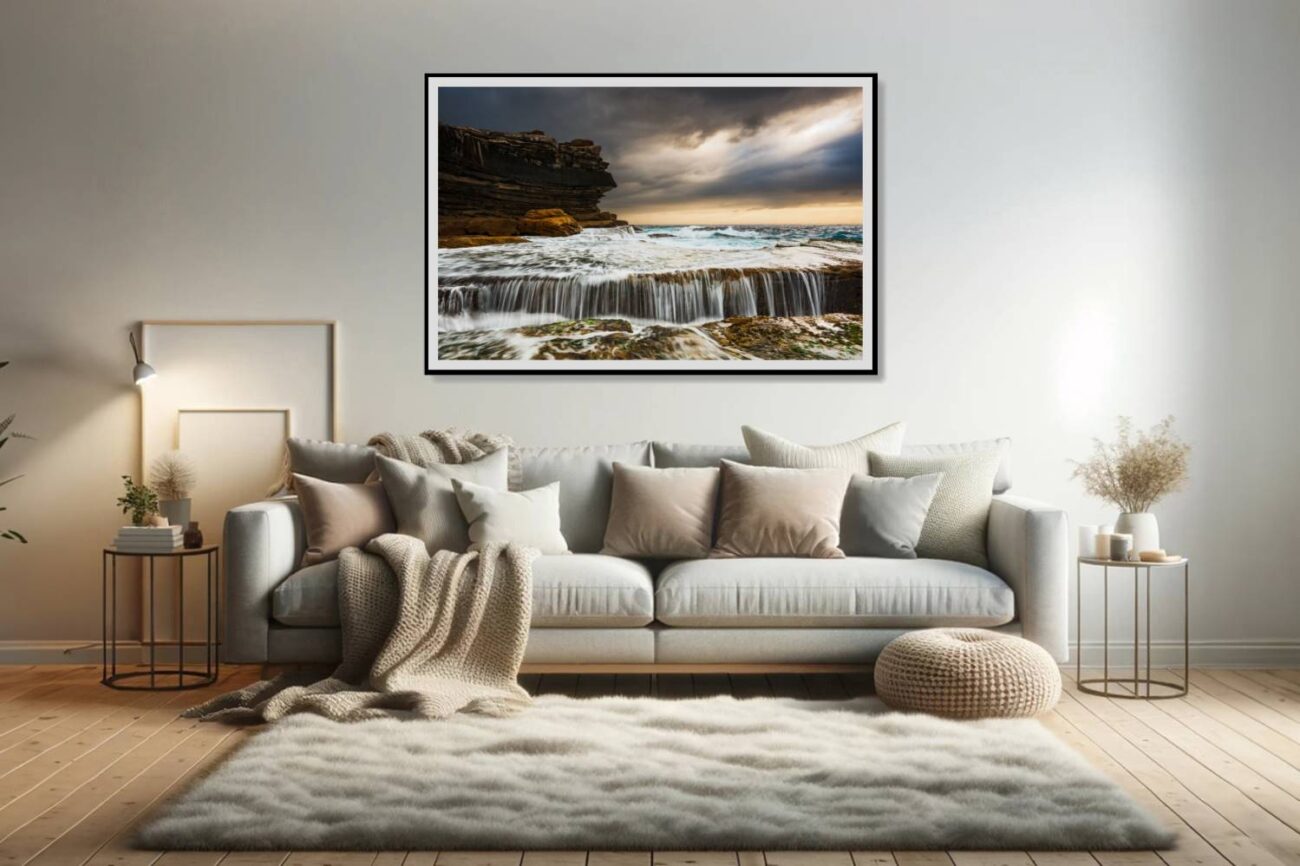 The living room features a framed art piece depicting a stormy sunrise at Clovelly Beach, where water cascades over rocky ledges into the abyss below, set against a tumultuous sky. This artwork adds a dramatic and dynamic element to the room, capturing the power and beauty of nature's forces.