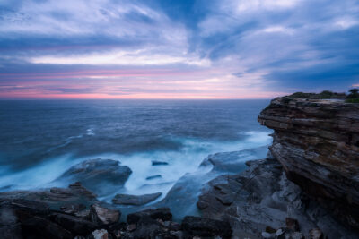 Sunrise gently illuminates Clovelly Beach, revealing soft waves and a serene sky in shades of pink and blue.