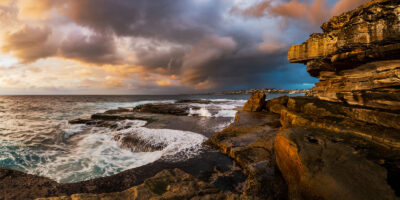 The sun casts a golden glow over turbulent waves and rocky shores at Clovelly Beach, under a storm-laden sky.