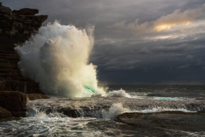 A massive wave crashes against the cliffs at Clovelly Beach during sunrise, with dark storm clouds overhead.