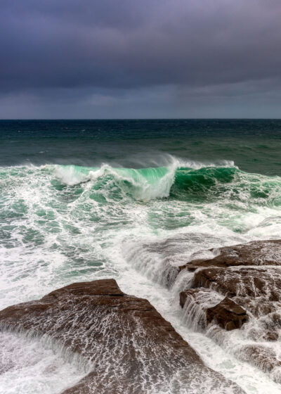 A large, turquoise wave gracefully bending as it approaches the rocky shore of Clovelly Beach under a subdued sky.
