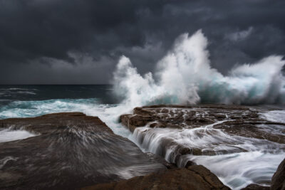 A tumultuous ocean storm seen at Clovelly Beach, with powerful waves erupting against the rugged coastline under a brooding sky.