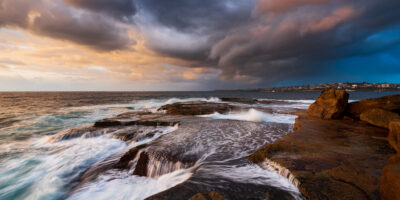Sunrise at Clovelly Beach under stormy skies with waves crashing, in an ocean print titled Indigo Fury.