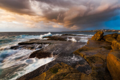 The first light of day peeks through storm clouds over Clovelly Beach, highlighting the dramatic interaction between the rugged shoreline and the churning ocean waves.