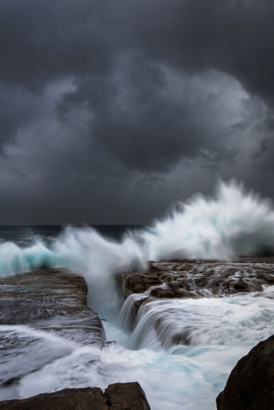 Giant waves at Clovelly Beach clash against the rocks, sending a dramatic spray into the air under a stormy sky.