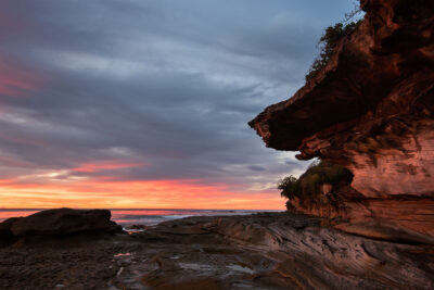 Soft morning light whispers over the textured cliffs of Cronulla, painting a serene pink and orange scene.
