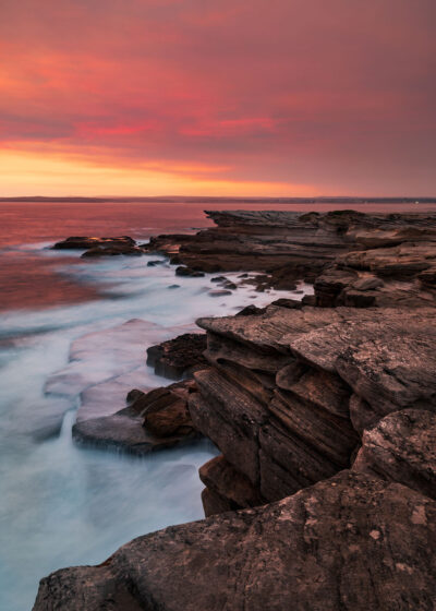 Waves crashing against Potter Point cliffs under a ruby-colored sunset sky.