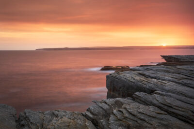 Sunset viewed from Potter Point, with a smoky sky and smooth sea reflecting fiery hues.