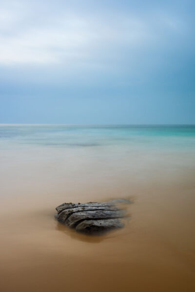 A solitary rock surrounded by the tranquil blue waters of Stanwell Beach, symbolizing seclusion in minimalist ocean art.