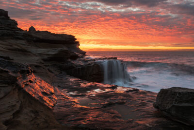 The cascading waters of Mahon Pool glow under the fiery sunrise in the vibrant coastal scene titled The Morning Ignition. Red wall art.