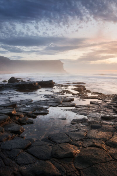 Sunrise casts a golden glow over the rocky shore of Garie Beach in a coastal photograph.