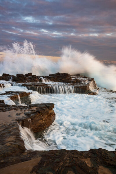 Waves crash against the rocks at Garie Beach in a dynamic display, saluting the day’s end with a roar in this sunset photo.