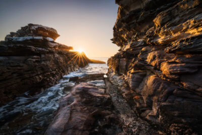 Sunrise casting an amber glow on textured cliffs with sea waves at Little Bay Beach.