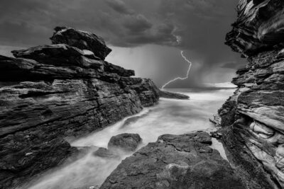 Black and white image of a stormy sea at Little Bay Beach, with lightning striking over tumultuous waters.