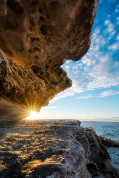 Sun casting a fiery gaze through an eroded rock formation against a blue sky at Little Bay Beach.
