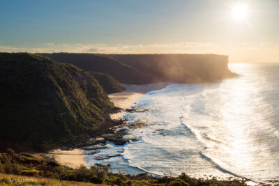 The sun rises over Little Garie Beach, casting a golden glow on the cliffs and waters in this peaceful morning photo.