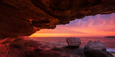 A spectrum of crimson colors the dawn sky, seen from a cave at Mahon Pool, in this vibrant and dynamic ocean sunrise art, panoramic wall art.