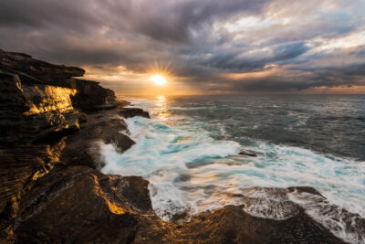 A dramatic Mahon Pool sunrise brings The Revival of day, with golden sunlight illuminating the cliffs and wild sea.