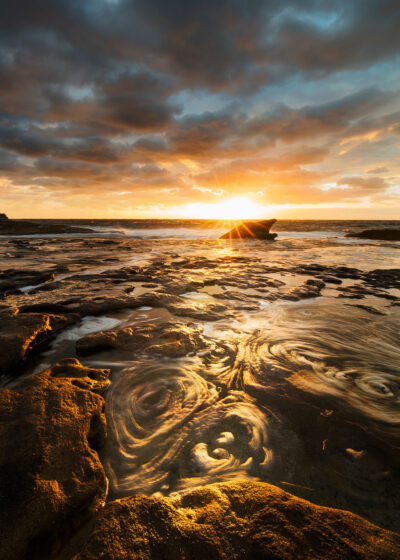 Rock pools at Mahon Pool reflecting the golden light of dawn in swirling patterns, captured in the serene photo Honey Swirls. Orange wall art.