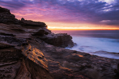 The rugged cliffs of Maroubra Ocean Pool are bathed in the purple hues of sunrise in the tranquil coastal photo The Enlightened Cliffs. Purple wall art.